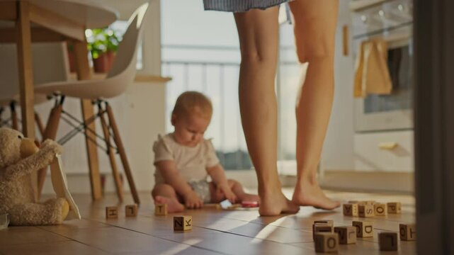 A young woman gracefully steps barefoot against the backdrop of a toddler sitting on the floor. The mother moves around the kitchen space while the child is nearby. Close-up on the woman's legs