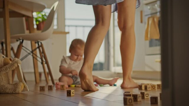 The legs of young woman are visible in the foreground as she walks around the house, while a small child plays on the floor. The mother slowly moves around the kitchen, with a child in the background
