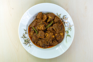 Delicious mutton curry in a white ceramic plate on a wooden background. Mutton bhuna is a popular, flavorful, and spicy Bangladeshi dish. Top view.