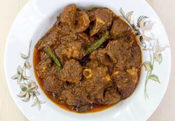 Close-up of spicy mutton curry in a white plate on a wooden background. Mutton bhuna is a popular, flavorful, and spicy Bangladeshi dish.