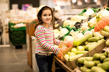 Kid little cute girl shopping for fresh organic fruits and vegetables in supermarket. Little girl holding pumpkin in a hands.