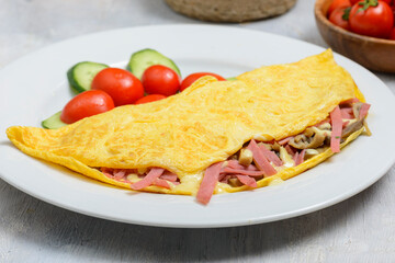 High angle studio shot of an omelette with salami, mushrooms and cheese on white plate with cherry tomatoes and cucumber slices on the side and a wooden bowl of cherry tomatoes in the background 