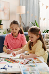 Two young girls engaging in drawing and coloring activities at home, sitting at a white table surrounded by colored pencils, crayons, and pictures, with cozy living room in background