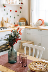 Holiday-themed dining table with a glass vase of red berries and pinecones on a red runner. Pink glasses and pie in the background, creating a warm, festive atmosphere in the kitchen