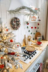 Festive kitchen setup with Christmas tree, pinecone wreath, and garland. Wooden countertops, white cabinets, pie on the stove, creating cozy holiday environment. View through a blurred Christmas tree