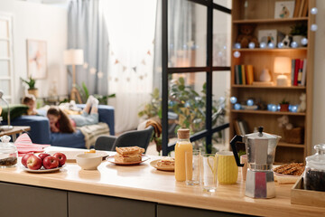 Kitchen counter with apples, orange juice, coffee maker, and pastries. People seen relaxing in background in a warmly lit, homey environment
