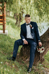 A man in a suit and tie sits on a tree stump with a bouquet of flowers in his hand