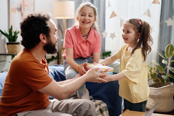 Smiling father handing colorful gift box to cheerful daughters in cozy living room, featuring...