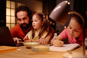 Father helping two daughters with homework at home. Scene showing family engagement, with laptop, books, and lamp on table creating a warm atmosphere