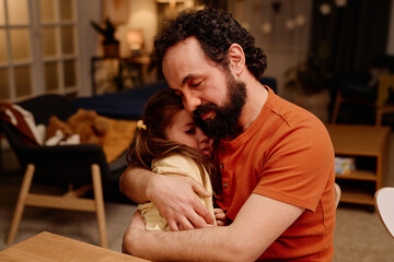 Warm scene showing father with beard gently hugging his young daughter at home, indicating love and comfort. Room in background filled with warm light and homely items