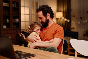 Father with beard and child cuddling at home desk in warmly lit room surrounded by cozy decor displaying strong emotional bond and tender moment