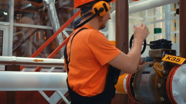 Closing gas pipeline industrial valve. Gas engineer in an orange jumpsuit and helmet operates valve while using walkie talkie, prioritizing safety and efficiency in industrial environment