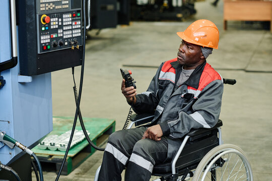 Worker in wheelchair operating industrial machine, wearing orange safety helmet and protective gear, ensuring safety and efficiency in factory environment