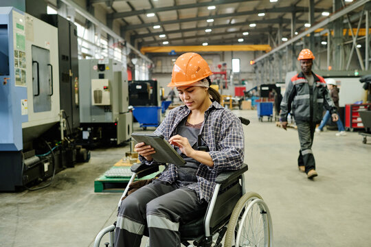 Worker in wheelchair using tablet for managing processes in modern industrial facility. Colleague in background walking between machines, both wearing safety gear - Powered by Adobe