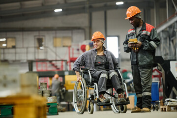 Wheelchair-using worker conversing with a colleague in an industrial warehouse setting, both...