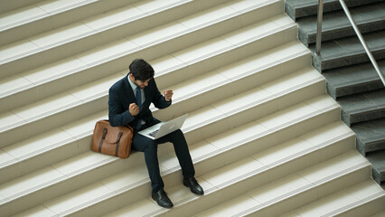 Top view business man celebrate successful project while sitting at stairs. Smart project manager getting new gob, getting promotion, increasing sales while calling friends by using laptop. Exultant.