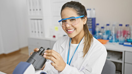 A cheerful hispanic woman scientist in a lab coat examines a sample with a microscope in a modern laboratory.