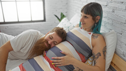 Couple enjoying a tender moment as man listens to baby's movements by resting his head on pregnant woman's belly while lying on a bed in a cozy bedroom.