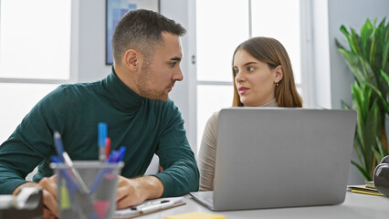 A man and woman engage as coworkers in an office, discussing over a laptop against an indoor plant backdrop.
