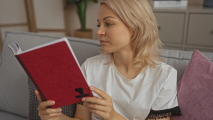 A young attractive blonde caucasian woman reading a red book indoors in a cozy living room with...