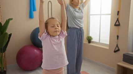 Mother and daughter exercising together indoors at a gymnasium, showcasing a family bonding moment with a woman and child practicing yoga in a sport center