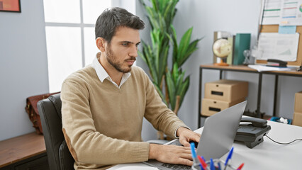 Handsome hispanic man working attentively on laptop in a modern office setting.