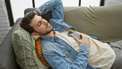 Fototapeta premium A young hispanic man reclining on a sofa with a smartphone in a cozy living room setting, embodying relaxed home life.