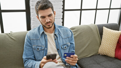 A young hispanic man with a beard sits in a modern living room using a smartphone and holding a credit card.