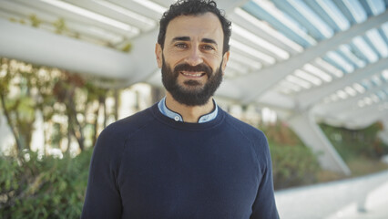 Handsome middle-aged bearded man smiling in a modern park with greenery and architectural structures.