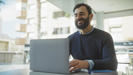Handsome middle-aged man with beard smiling while working on laptop in modern office setting.