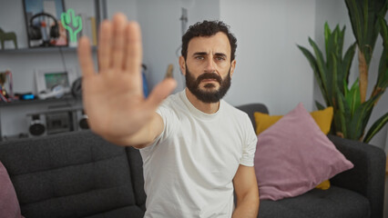 A serious bearded man gestures a stop sign with his hand in a modern living room setting,...