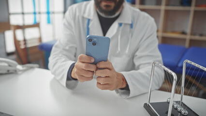 A bearded doctor holding a smartphone in a modern hospital office, with a stethoscope and newton's...