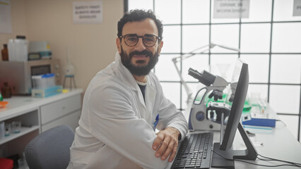 A smiling bearded man in a lab coat sitting at a desk in front of a computer inside a laboratory with equipment.