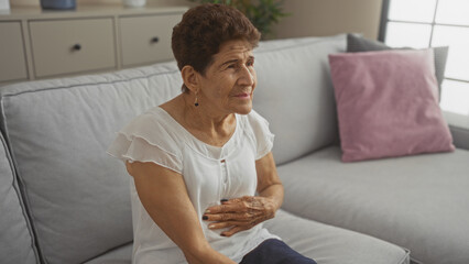 Elderly hispanic woman with short hair sitting in a living room, looking pensive and touching her stomach, in a cozy home setting.