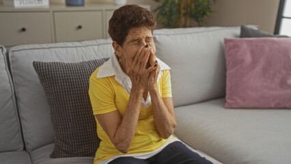 An elderly hispanic woman with short hair sits in a living room, covering her face with her hands on a couch with pillows in a cozy home interior.