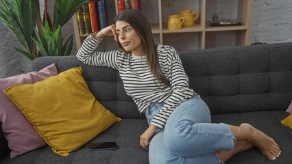 Obraz premium A thoughtful young hispanic woman lounges casually at home on a gray sofa surrounded by colorful cushions and a cozy interior.