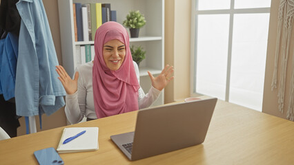 A smiling woman in a pink hijab engaging in a video call on a laptop at a home office setting