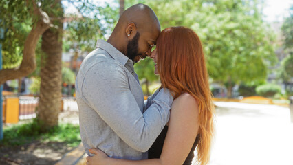Fototapeta premium Interracial couple embracing in an urban park, man and woman smiling at each other with trees and city in the background, showcasing love and relationship outdoors