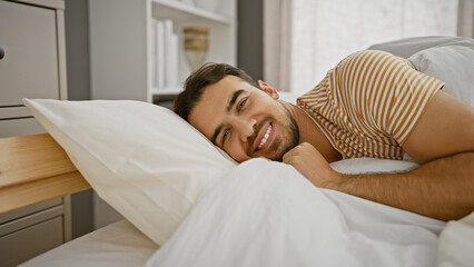 A handsome young hispanic man with a beard smiles while lying in bed in a cozy bedroom.