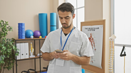 Hispanic male healthcare worker using smartphone in bright rehab clinic interior with exercise equipment.