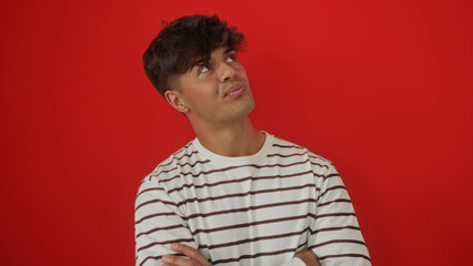 A handsome young hispanic man in a striped shirt crosses his arms and looks upwards against a vivid...
