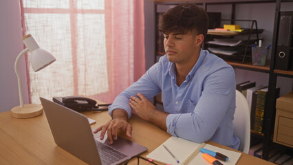 A handsome young hispanic man in an office setting, working on a laptop while surrounded by office supplies and furniture in a well-lit room.