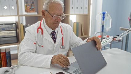 Obraz premium Senior caucasian man doctor in a clinic using a laptop in a hospital room.