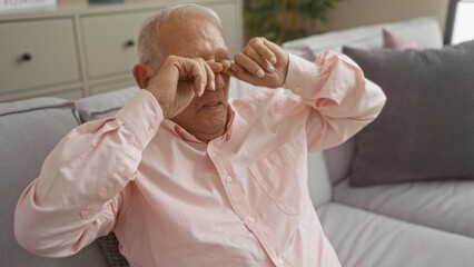 Elderly handsome caucasian man in a pink shirt sits tired and rubbing his eyes while relaxing in his home living room, showcasing a mature, grey-haired senior in a cozy indoor apartment setting.