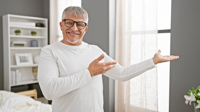 A cheerful middle-aged man in a white shirt presenting something in a bright bedroom with a welcoming gesture.