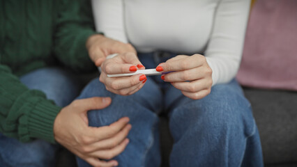 A man and woman sit closely, anxiously awaiting the result on a pregnancy test in a cozy living room.