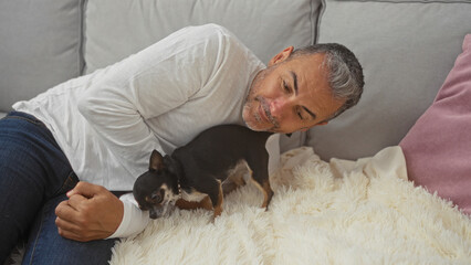 Middle-aged hispanic man lying on a couch with his chihuahua dog in a cozy living room.