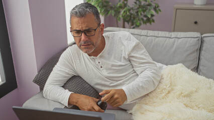 Attractive middle-aged hispanic man with glasses working on a laptop while relaxing on a cozy living room sofa in his apartment, wearing casual clothes and holding a remote control.