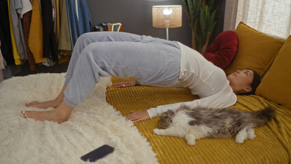 A young asian woman exercises on her bed next to her pet cat in a cozy bedroom interior with soft lighting.