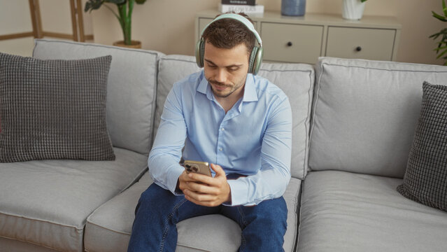 Handsome young hispanic man with a beard wearing headphones and sitting on a couch in a bright living room using a smartphone immersed in his home surroundings.